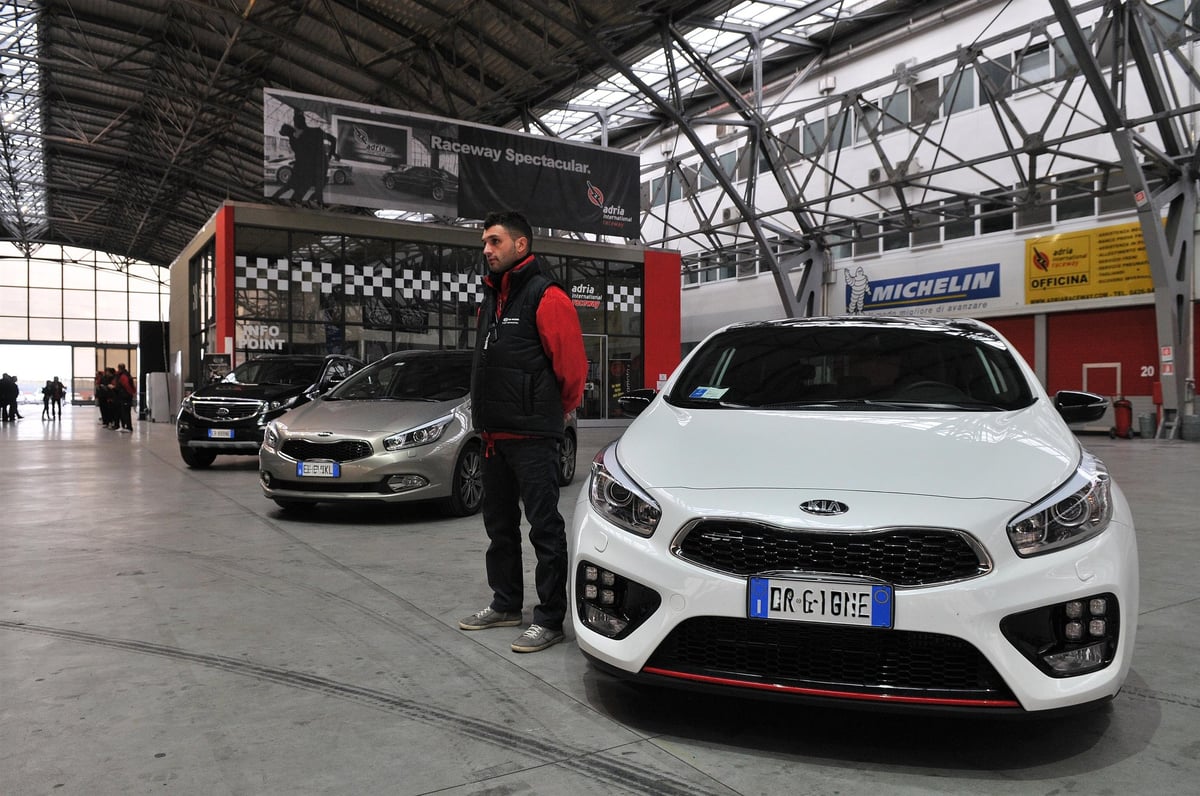 Man standing beside white Kia sports car at indoor auto show or exhibition hall with metal framework