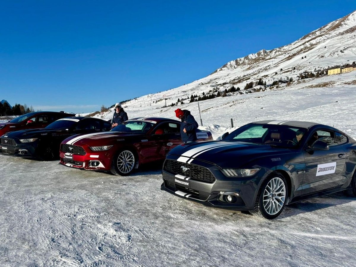 Performance cars lined up on a snowy mountain slope with spectators and snow-covered peaks in the background